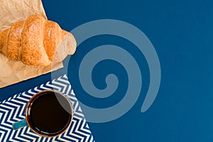 Top view of cup of black coffe and croissant on craft paper on blue background with copy space. Morning breakfast in french style