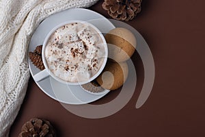 Top view of cup of cocoa with marshmallows, cookies, white sweater and pine cones on brown background