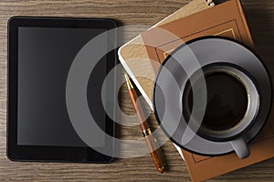 Top view of cup of coffee, blank screen of tablet pc, book, notebook ad pen on the wooden desk.Empty space