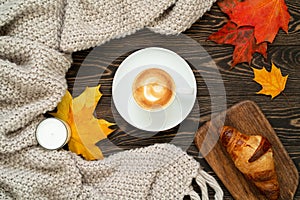 Top view of a cup of coffee, fresh croissant, candle, colorful maple leaves and knitted plaid on the wooden table. Autumn flat lay