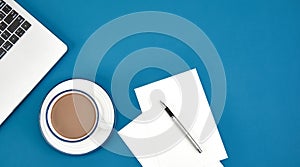 Top view of a cup of coffee, laptop, papers, and a pen on a blue surface