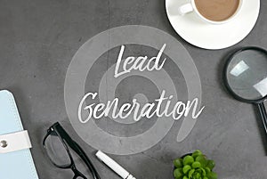 Top view of a cup of coffee, magnifying glass, plant, pen,sunglasses and notebook on grey floor written with Lead Generation photo