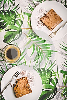 Top view of a cup of coffee and two dessert plates with brownie cake over table-cloth with green leaves pattern