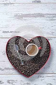 Top view of a cup of espresso in a red cup on top of freshly roasted coffee beans in a heart shaped tray on textured white wooden