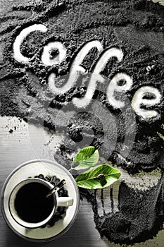 Top view on cup of hot black coffee with green leaves and text word hand written in english `coffee` in milled coffee texture