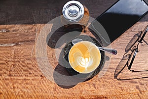 top view cup of hot milk coffee with latte art, smartphone, eye glasses and brown sugar on wooden desk in the morning light