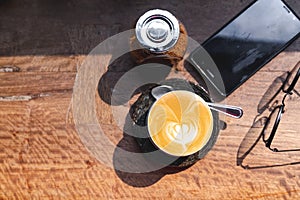 top view cup of hot milk coffee with latte art, smartphone, eye glasses and brown sugar on wooden desk in the morning light