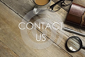 Top view of a cup of milk tea,eyeglasses,magnifying glass,notebook and pen on wooden background written with CONTACT US