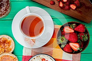 top view of cup of tea with white chocolate on teabag and bowl of strawberries with crispbreads and peach jam on green background