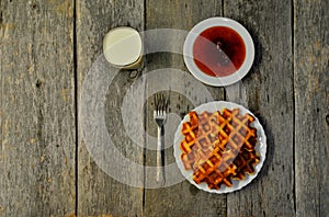 Top view of curd waffles on a plate, apricot jam, a glass of milk and a fork on a wooden table