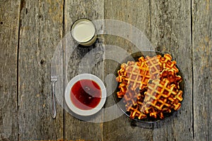 Top view of curd waffles on a plate, apricot jam, a mug of milk and a fork on an old wooden table