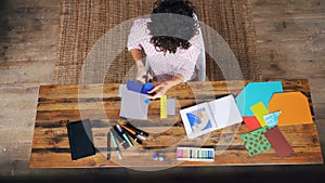 Top view of curly-headed woman making colorful paper collage sitting at table cutting figures with scissors. Design