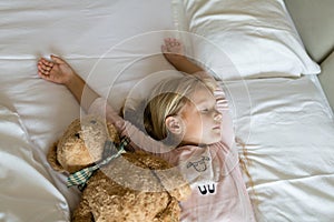 Top view of cute blonde little girl in pajamas lying in white bed with teddy bear, awaking early in the morning before going to
