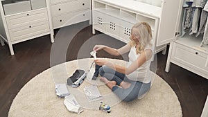 A top view of a cute pregnant blonde sitting on the carpet. She folding baby`s T-shirts