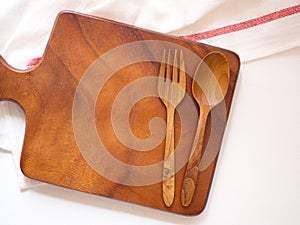 Top view, cutting board with wooden fork and spoon, fabric or napkin over white wooden background.
