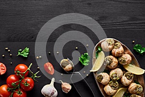 Top view of delicious cooked escargots on black wooden table with ingredients and tweezers.