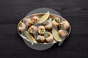 Top view of delicious cooked escargots with lemon on black wooden table.