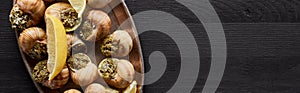 Top view of delicious cooked escargots with lemon on black wooden table, panoramic shot.