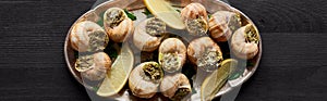 Top view of delicious cooked escargots with lemon on black wooden table, panoramic shot.