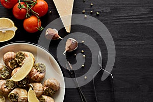 Top view of delicious cooked escargots with lemon slices on black wooden table with ingredients.