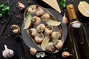 Top view of delicious cooked escargots with lemon slices on black wooden table with spices, Parmesan and white wine.
