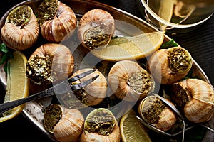 Top view of delicious cooked escargots with lemon slices and cutlery on plate on black wooden table.