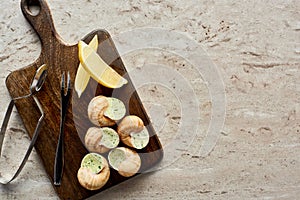 Top view of delicious cooked escargots served on wooden cutting board with lemon slices and tweezers on stone background