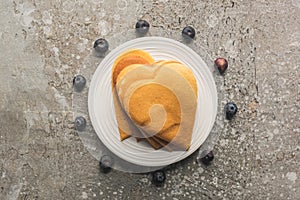Top view of delicious heart shaped pancakes on plate near blueberries on grey concrete surface