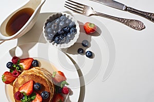 Top view of delicious pancakes with blueberries and strawberries on plate near cutlery and maple syrup in gravy boat on marble