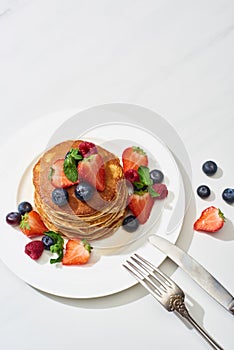 Top view of delicious pancakes with maple syrup, blueberries and strawberries on plate near fork and knife on marble white surface