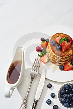 Top view of delicious pancakes with maple syrup, blueberries and strawberries on plate near fork and knife on marble white surface