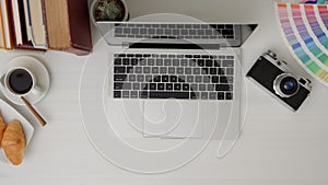 Top view of  designer workspace with laptop, camera, colour swatch, breakfast and decorations on white desk