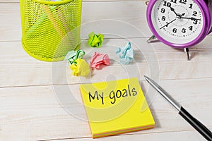 Top view of a desk with a clock and stickers and crumpled paper for writing. My goals are written on a yellow sticker