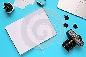 Top view of a desktop of a photographer consisting on a camera, a laptop, a notebook and a memory card on a blue desk background -