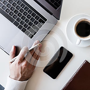 Top view of the desktop, on which there is a cup of coffee, a laptop and a notebook for records. Man freelancer works at a table