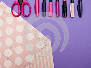 Top view of different pink and purpule pens or markers and scissors on purple and pink paper background with space for text