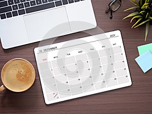 Top view, digital tablet PC with calendar app show December\'2023 month screen and laptop, coffee cup on brown workplace.