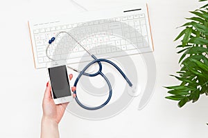 Top view doctor`s hand holding smartphone, stethoscope and keyboard on white table.