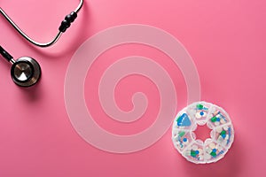 Top view daily doses pills in plastic container and a stethoscope on pink background