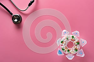 Top view daily doses pills in plastic container and a stethoscope on pink background