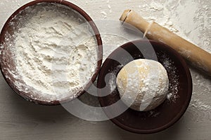 Top view of a dough on a plate, a bowl full of flour, and a rolling pin