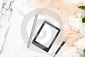 Top view e-book reader, paper notepad, white peony flowers, pastel color scarf on marble background. Minimal flat lay style