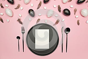 Top view of easter eggs, feathers on pink background. Table setting, black plate and cutlery, white card.