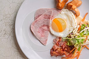Top view easy breakfast menu, fried egg, ham, bacon, sausage, and salad. Breakfast on white plate on white stone table, natural