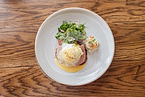 Top view of Eggs Benedict with ham, toast and mashed potato. Served with salad in white plate on wooden table