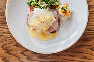 Top view of Eggs Benedict with ham, toast and mashed potato. Served with salad on a white plate on wooden table