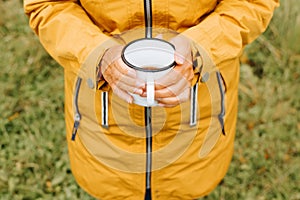 Top view of elderly woman's hands are holding hiking iron mug with hot tea outdoors.