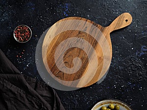 Top view of empty round wooden cutting board with handle, red pepper and green olives on abstract dark background