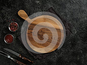 Top view of empty round wooden cutting board with handle, red pepper, sauce, meat fork and knife on beautiful dark background