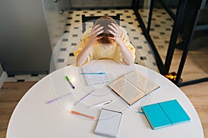 Top view of exhausted pupil boy tired from studying holding head head with hands sitting at desk with paper copybook